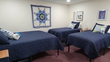 A room with three beds, each with a blue quilted bedspread and pillows, a red carpet, and wall art including a star pattern and two framed pictures.