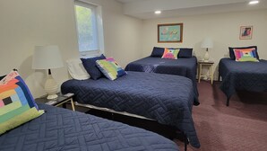 A hotel room with three beds, each with a blue quilted bedspread and colorful pillows. There is a window with blinds, a wall-mounted world map, and a lamp on a bedside table.
