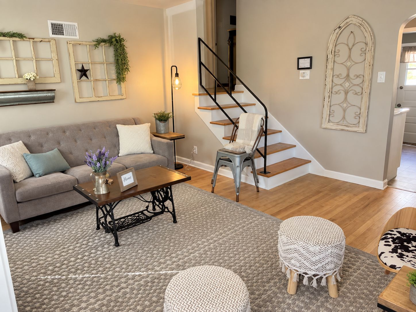 Sala de estar con sofá, mesa de centro y otomanas, escalera con una silla y elementos decorativos en la pared.