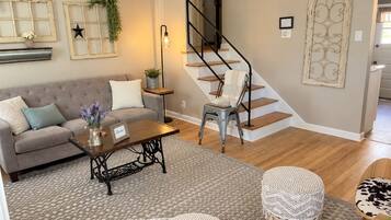 Sala de estar con sofá, mesa de centro y otomanas, escalera con una silla y elementos decorativos en la pared.