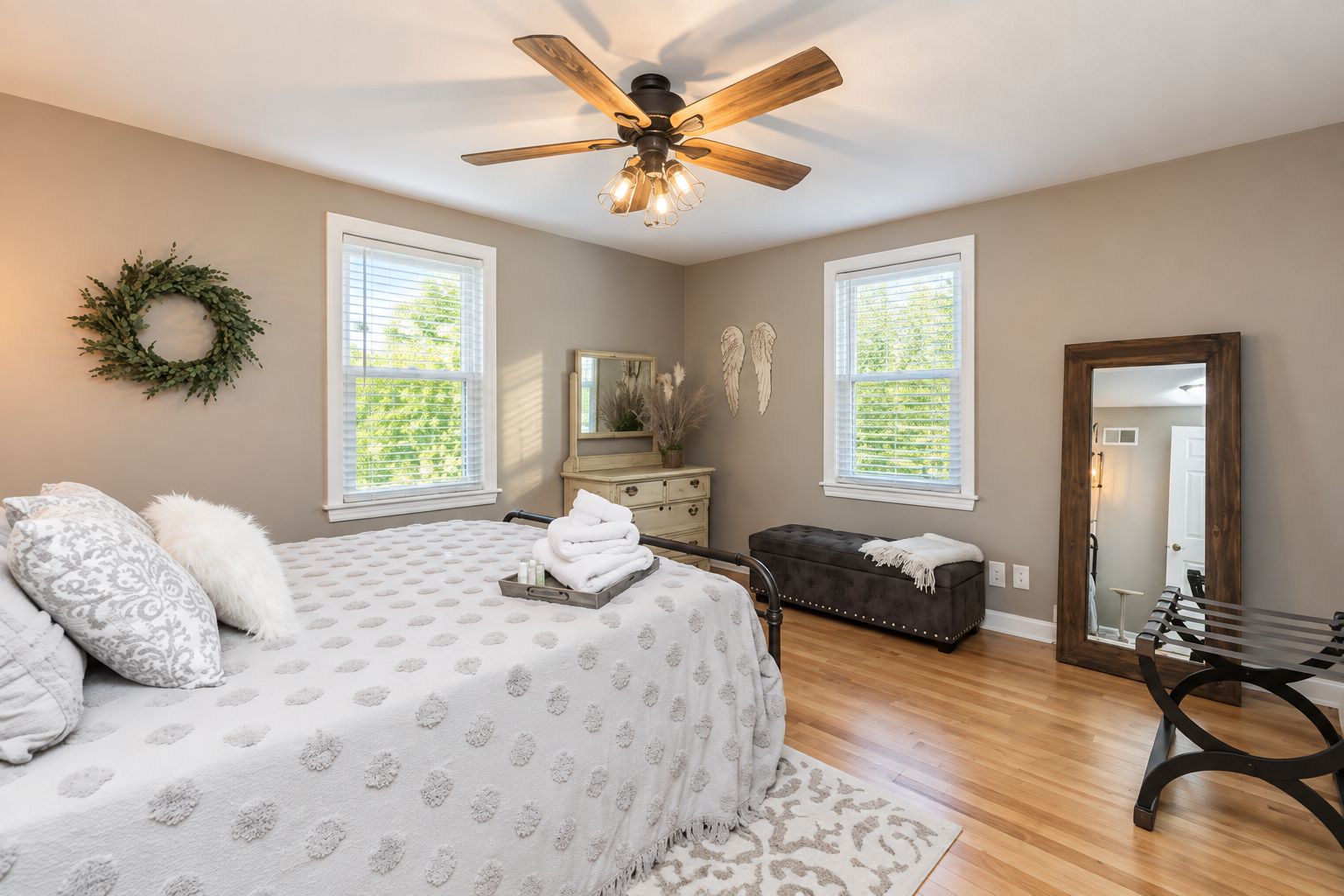 Une chambre à coucher comprenant un lit, une commode, un banc, un ventilateur de plafond et deux fenêtres.