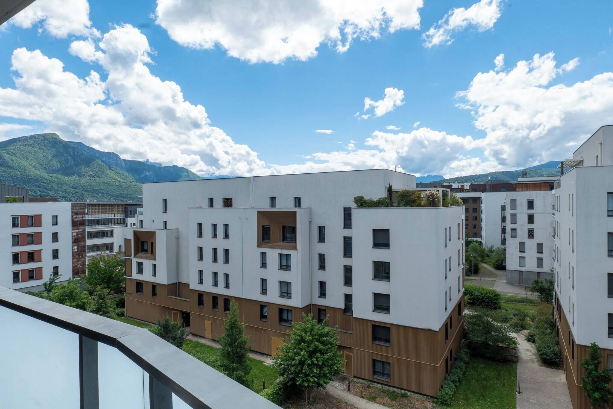 A modern residential building complex with multiple apartment buildings, a rooftop garden, and a mountainous backdrop.