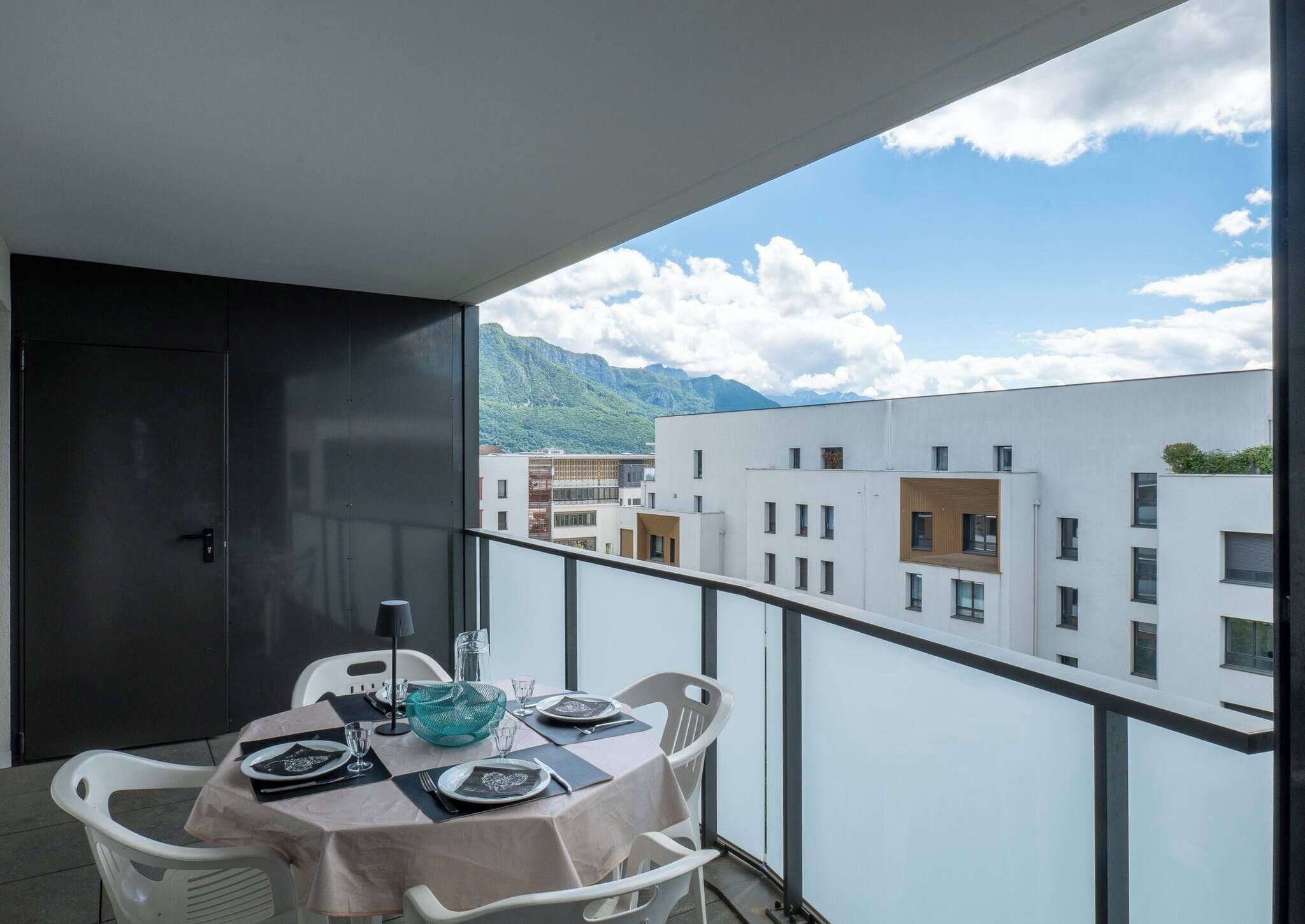 A balcony with a table set for dining, overlooking a mountainous landscape and urban buildings.