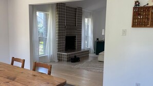 Een eetruimte met een houten tafel en stoelen, een open haard met betegelde omlijsting, en zicht op een woonkamer via een open doorgang.