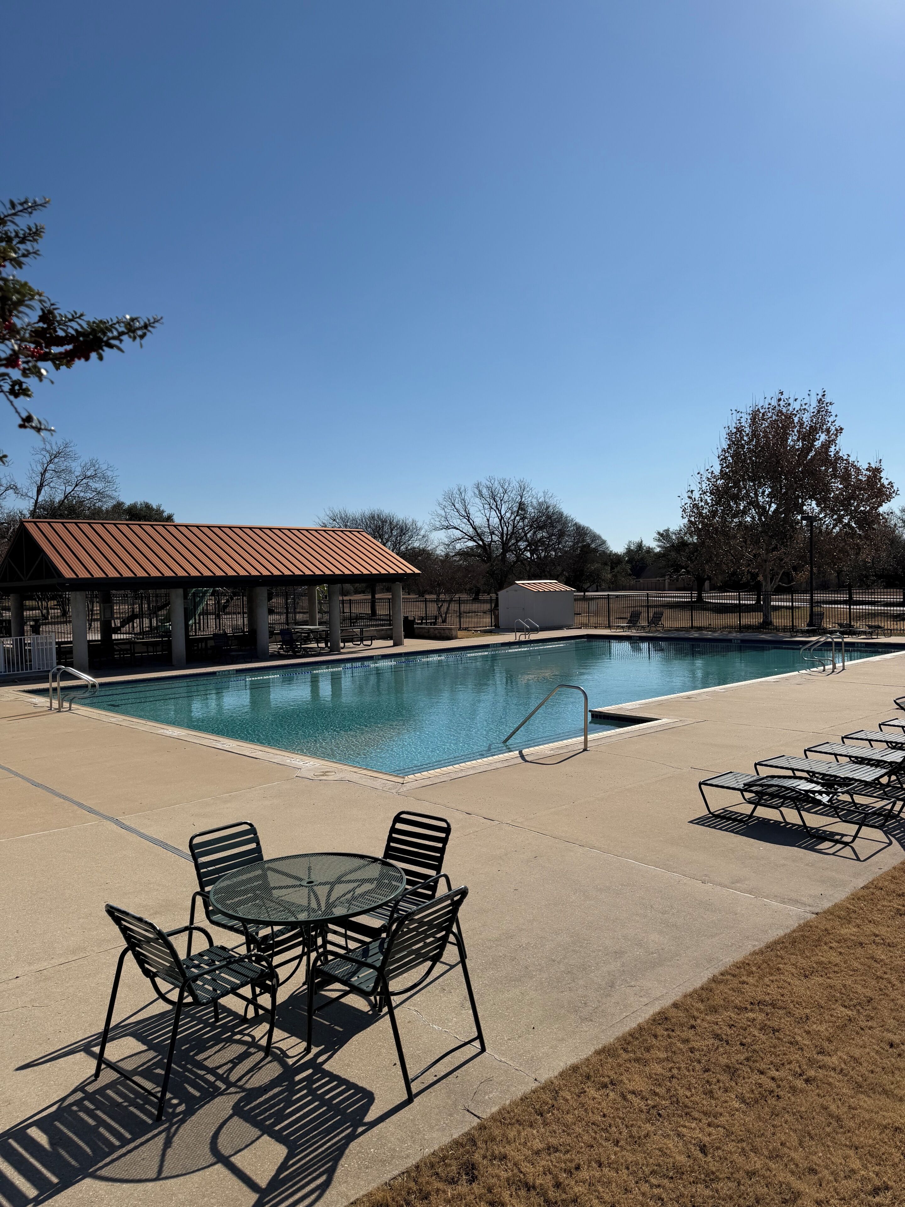 A swimming pool with a metal roof structure, lounge chairs, and a table with chairs.