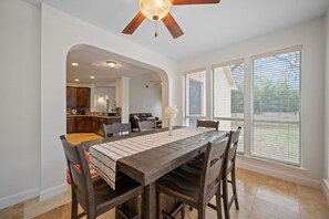 Uma sala de jantar com mesa e cadeiras de madeira, ventilador de teto e grandes janelas.