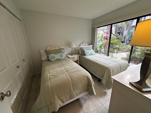 A bedroom with two beds, a nightstand, a lamp, and a window with a view of greenery. - Spacious two bedroom condo across the street from Siesta Key Beach (Sarasota)
