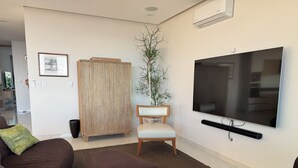 Een woonkamer met een grote flatscreen televisie, een bruine bank, een houten stoel en een potplant.