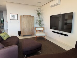 A living room with a large flat-screen TV, a brown sofa, a wooden chair, and a potted plant. - Fabulous Penthouse with Ocean and Mountain Views (Puerto Vallarta)
