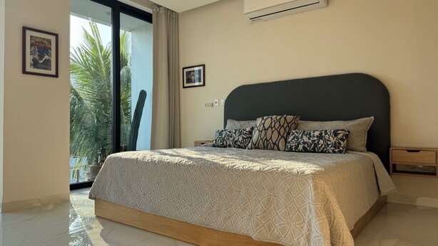 Een slaapkamer met een bed, nachtkastje, airconditioning en een groot raam met uitzicht op palmbomen.