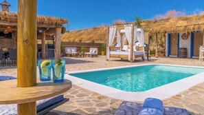 Zona junto a la piscina con terraza de madera, estructura de techo de paja, cama con dosel, sillones de descanso y una mesa con bebidas.