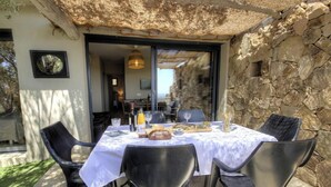 An outdoor dining area with a stone wall, a table set with food and drinks, and a thatched roof overhead. - Alba - Porto Vecchio (PORTO VECCHIO)