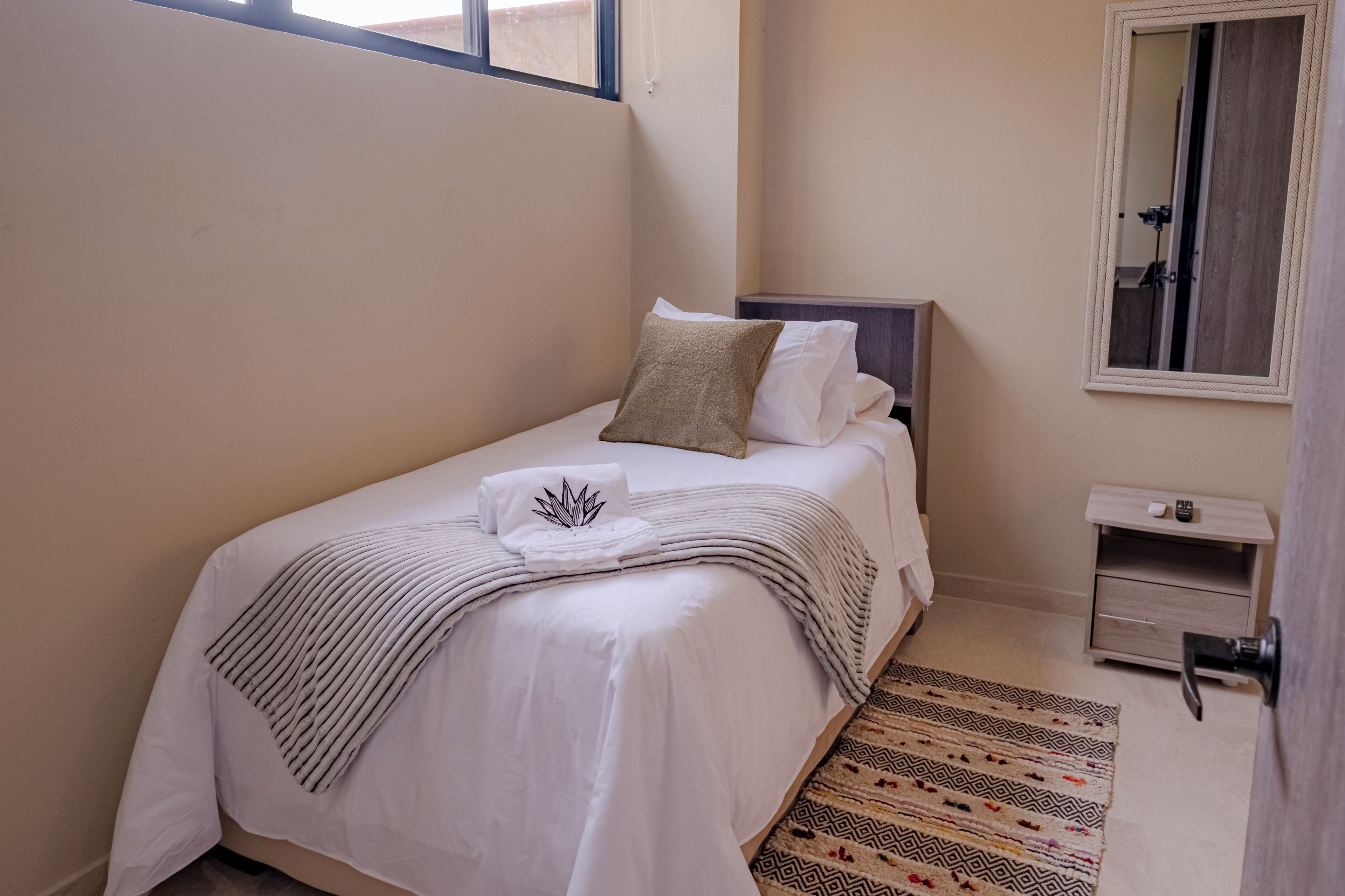 A neatly made bed with a white comforter and a decorative pillow, a small bedside table with a lamp, and a mirror on the wall.