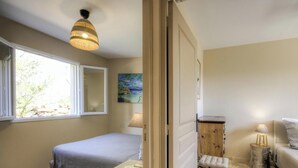 Quarto com cama, janela, quadro, abajur e um pequeno banquinho de madeira.