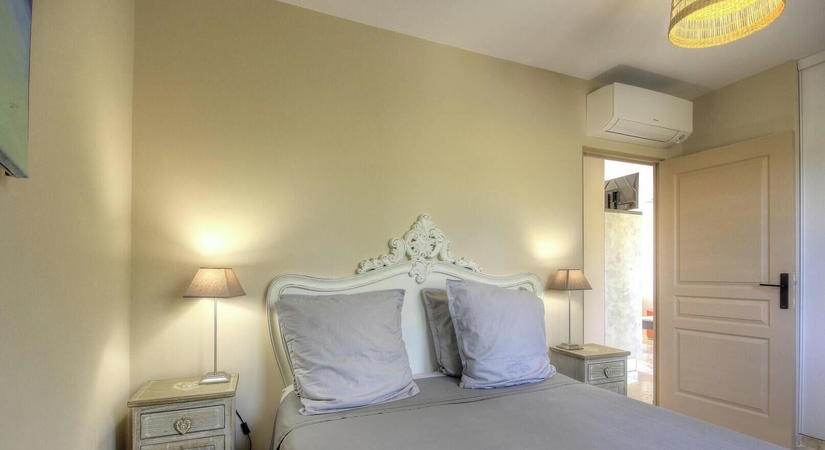 A bedroom with a bed, two bedside lamps, a nightstand, a wardrobe, and an air conditioning unit.