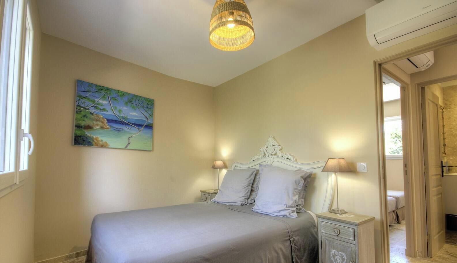 A bedroom with a bed, bedside lamps, a painting on the wall, and a door leading to another room.