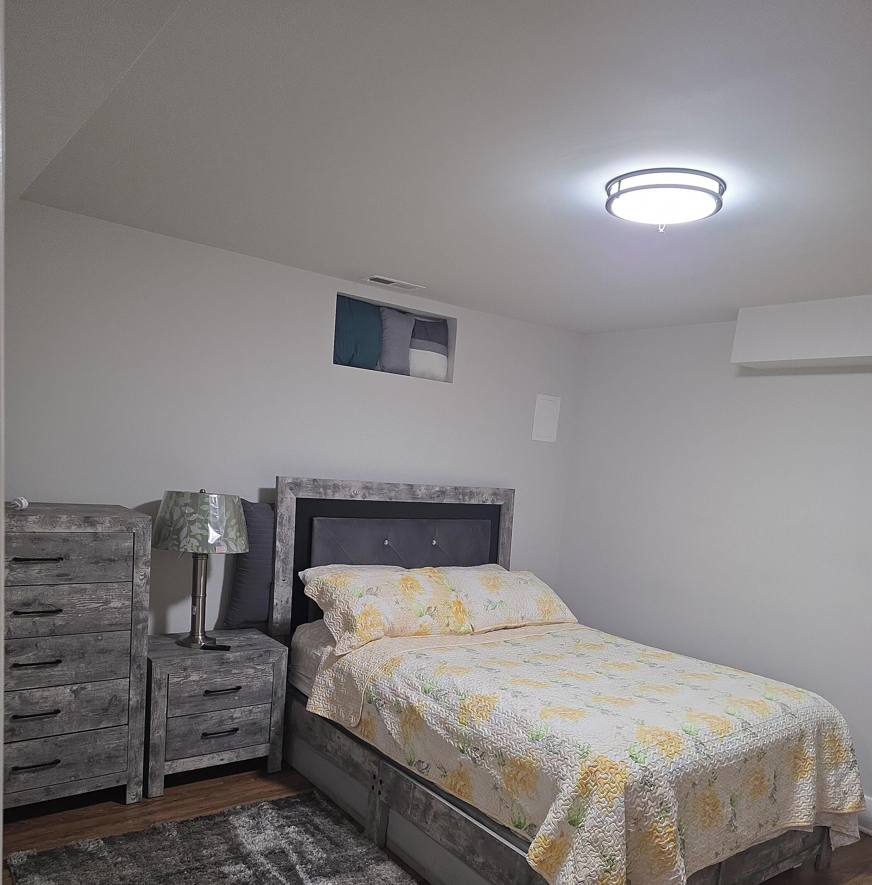 Et soveværelse med seng, natbord, lampe, kommode og loftlampe.