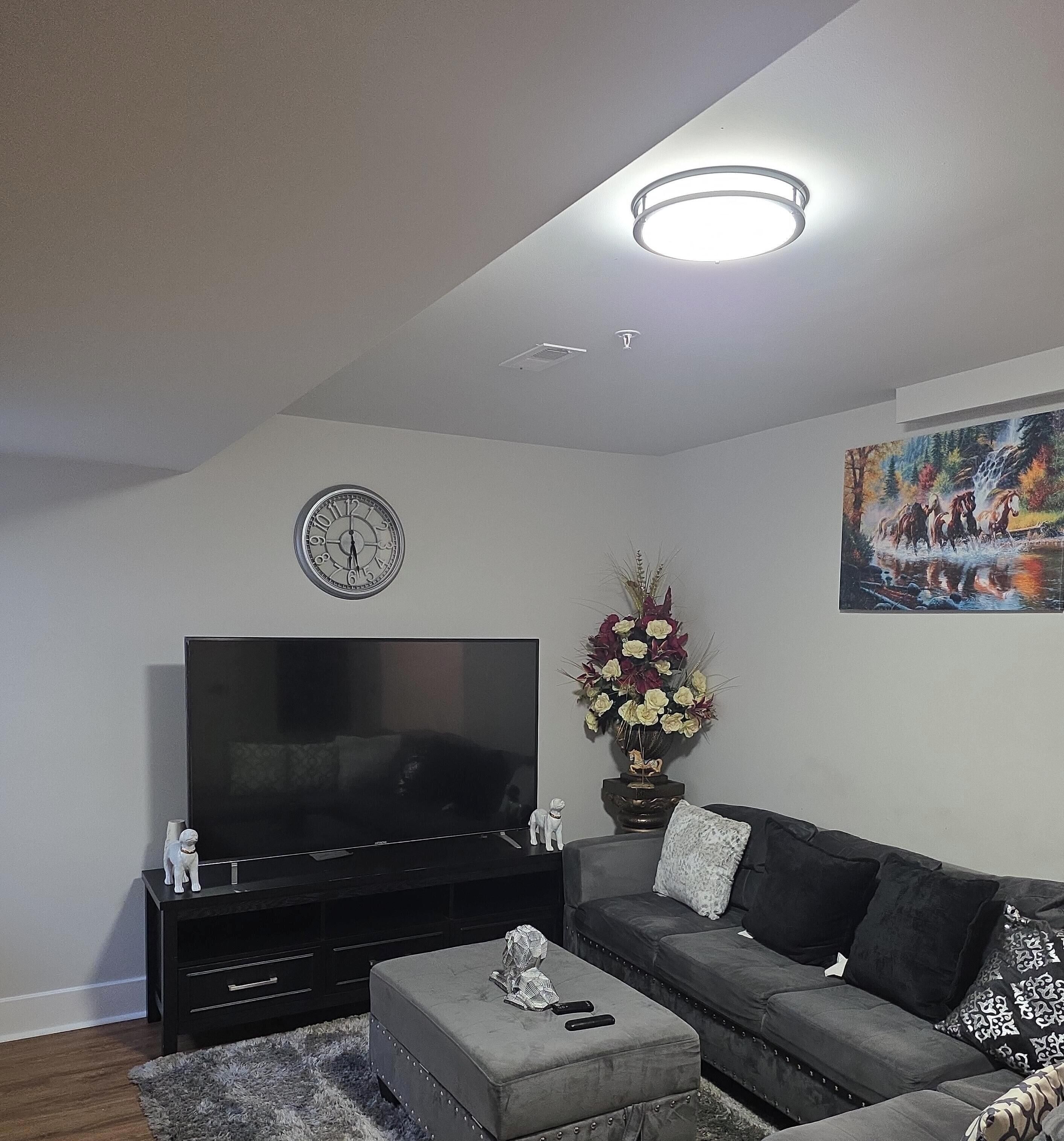 A living room with a flat-screen TV, a sofa, an ottoman, a clock, and a painting on the wall.