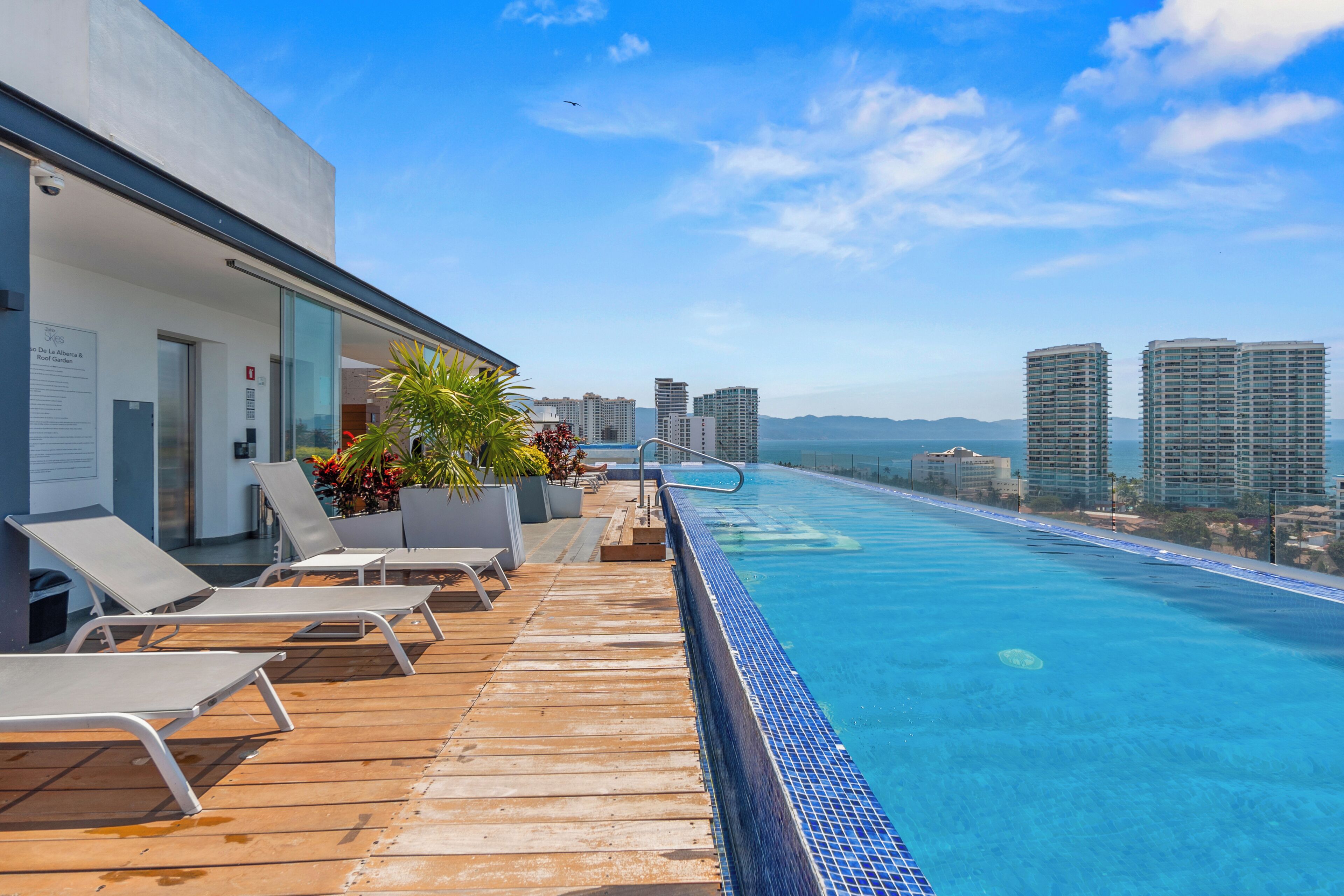 Ein Pool mit unendlicher Optik auf dem Dach, ausgestattet mit Liegestühlen, einem Holzterrasse und einer Stadtkulisse im Hintergrund.