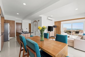 A modern apartment with a kitchen, dining area, and living room. It features a large window with a view of the city and a balcony. - Luxury Dual Condos at Zoho Skies Rooftop Pool (Puerto Vallarta)