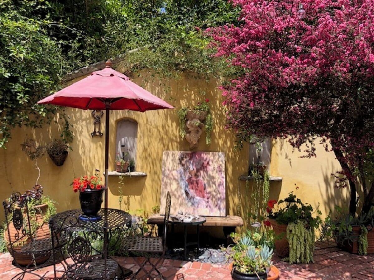Eine Terrasse im Freien mit einem roten Sonnenschirm, einem Tisch, Stühlen, Kübelpflanzen und einer Wand mit Wandmalerei.