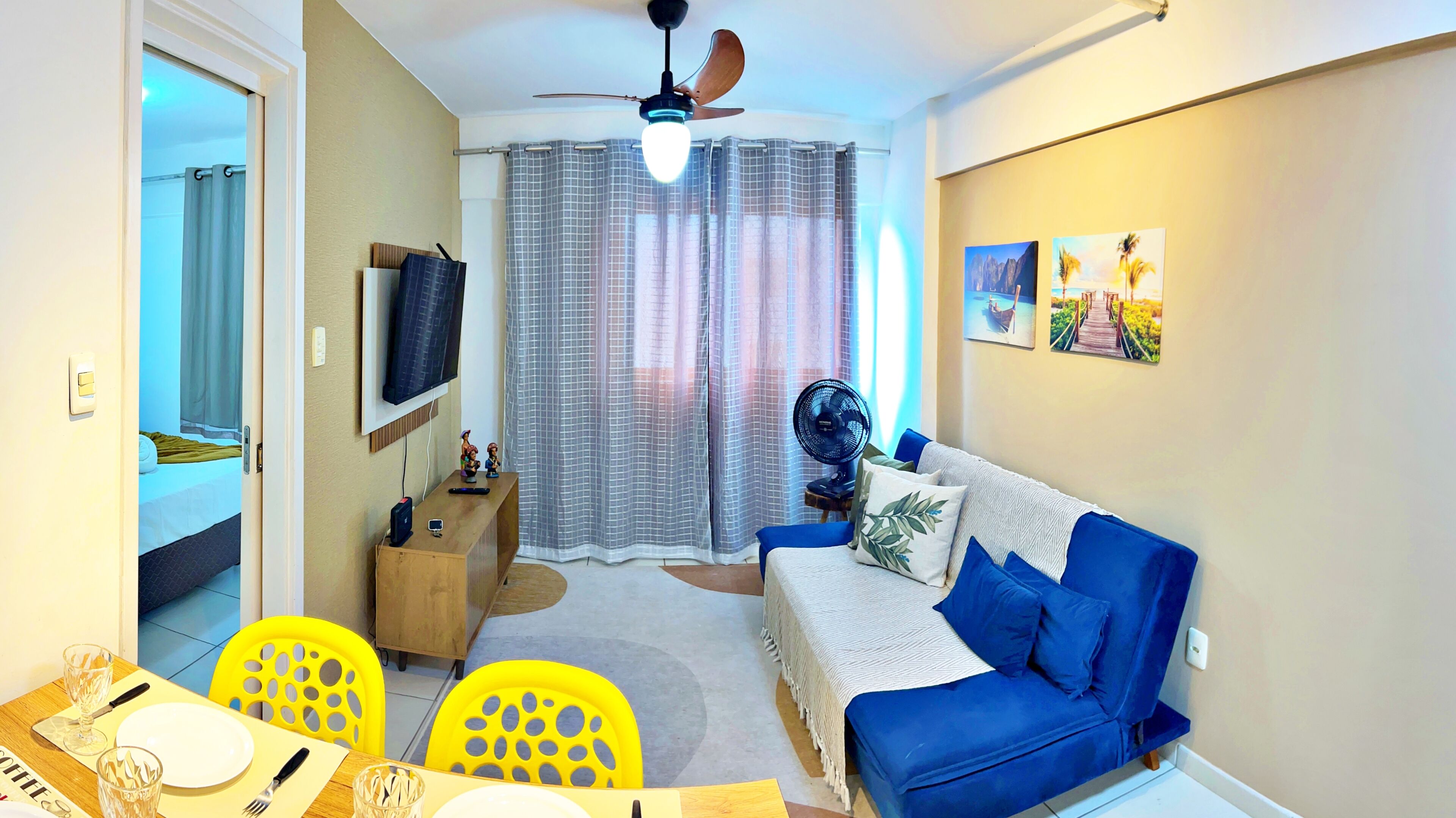 Uma sala de estar com sofá, televisão fixada na parede, ventilador de teto e um espaço de jantar com mesa e cadeiras.