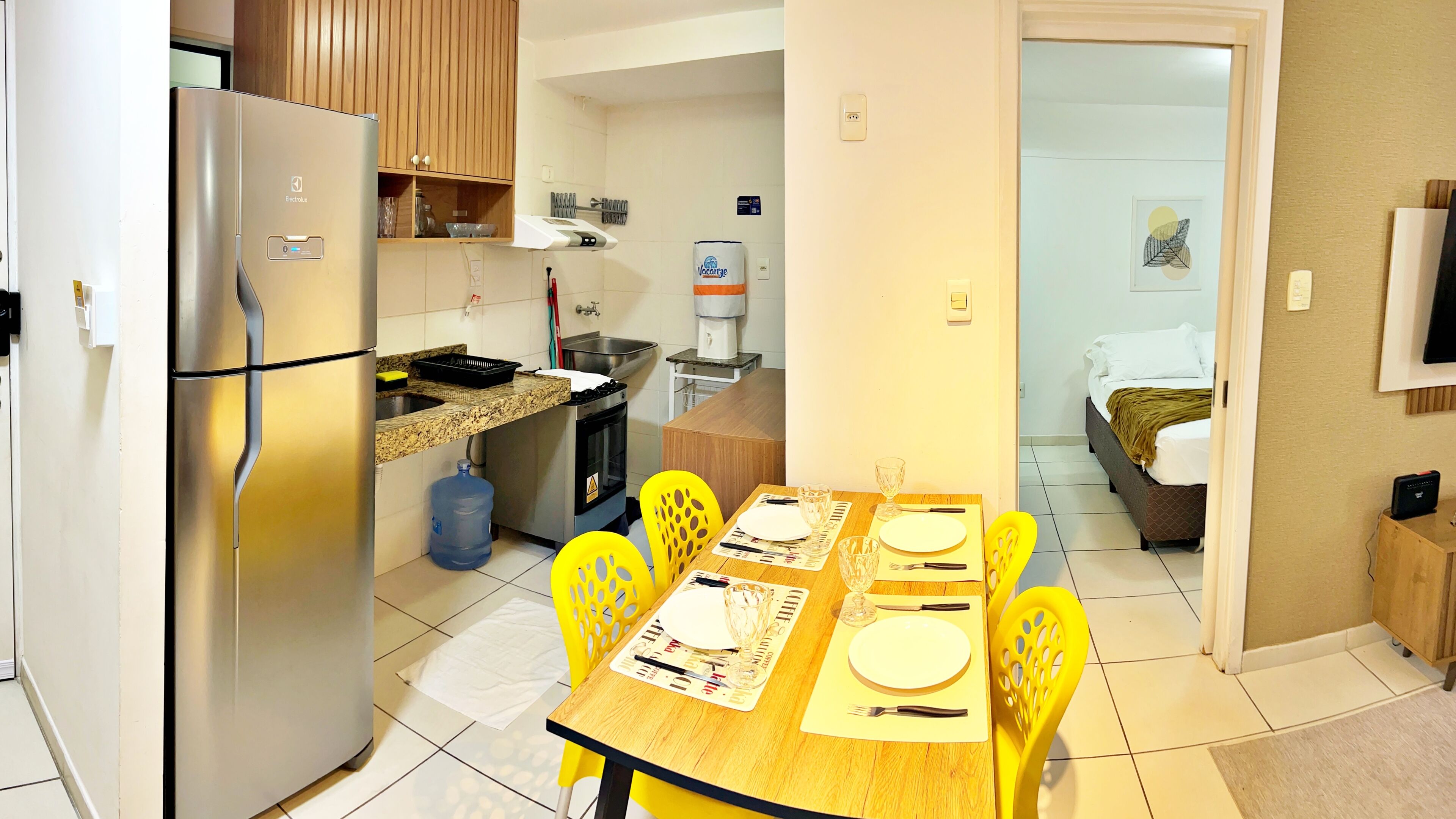 Cozinha compacta com geladeira, máquina de café e pia. Há uma área de refeições com uma mesa posta para dois, cadeiras amarelas e uma cama no quarto adjacente.