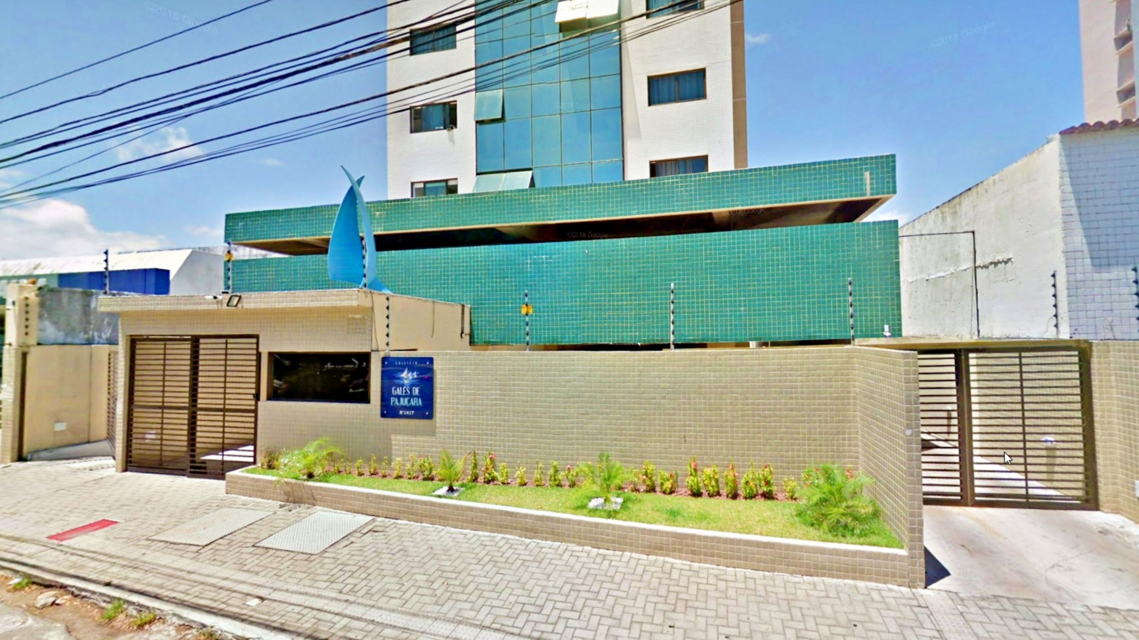 Um edifício moderno com fachada revestida de azulejos, uma pequena área ajardinada com plantas e uma placa com o nome 'Gales de Palacios'.