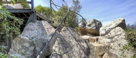 Una escalera de metal que asciende por un sendero rocoso, rodeada de grandes rocas y follaje.