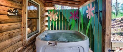 Una cabaña de madera con jacuzzi, una ventana y una cerca verde adornada con flores.