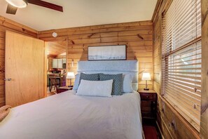 A wooden-paneled bedroom with a bed, bedside lamps, and a window with blinds. - Cozy Sevierville Cabin - Hot Tub & Fireplace (Sevierville)