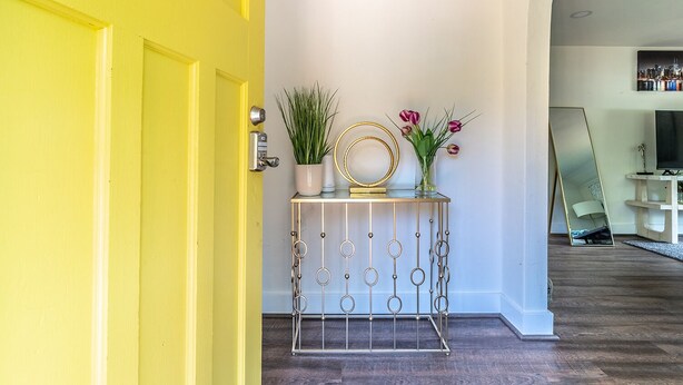 Een felle gele deur die leidt naar een hal met een kleine tafel waarop een plant, een bloevas en een decoratieve spiegel staan.