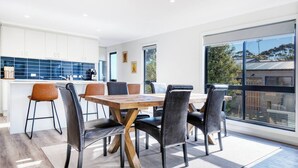 Un'area pranzo moderna con un tavolo e sedie in legno, una cucina sullo sfondo e ampie finestre che offrono vista all'esterno.