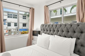 Ein Hotelzimmer mit einem großen Bett, weißer Bettwäsche, Blick ins Freie und einem mit Vorhängen versehenen Fenster.