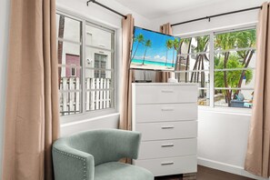 A room with a television on a stand, a white dresser, a green chair, and a window with a view of palm trees and a beach. - Adorable 1-bedroom apartment with Pool in Miami Beach (Miami Beach)