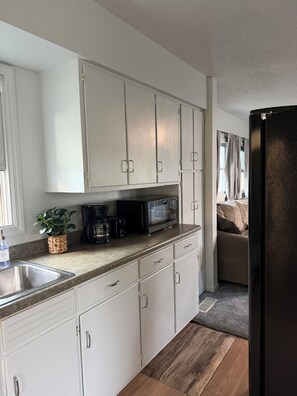 Cocina compacta con armarios blancos, pileta de acero inoxidable, microondas y cafetera.
