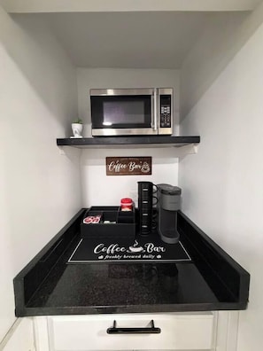 Eine Küche mit schwarzer Arbeitsplatte, einer Kaffeemaschine, einer Mikrowelle und einem Schild mit der Aufschrift 'Coffee Bar – frisch gebrüht täglich'.