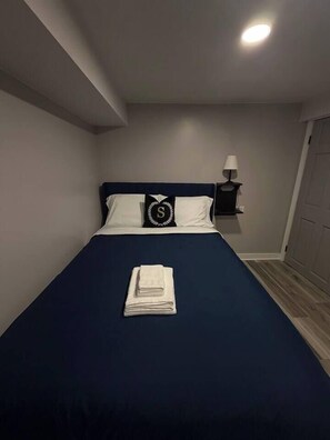 Een net opgemaakt bed met een donkerblauwe deken en witte kussens, een opgevouwen witte handdoek op het bed, en een nachtkastje met een lamp aan de rechterkant.