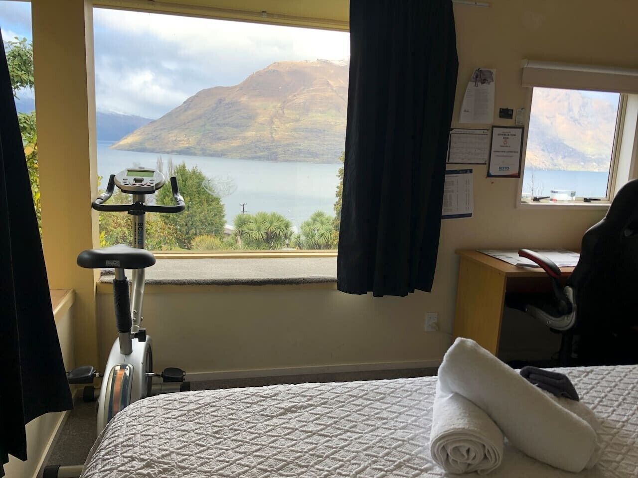 A hotel room with a bed, a desk, a chair, a treadmill, and a window with a mountain view.