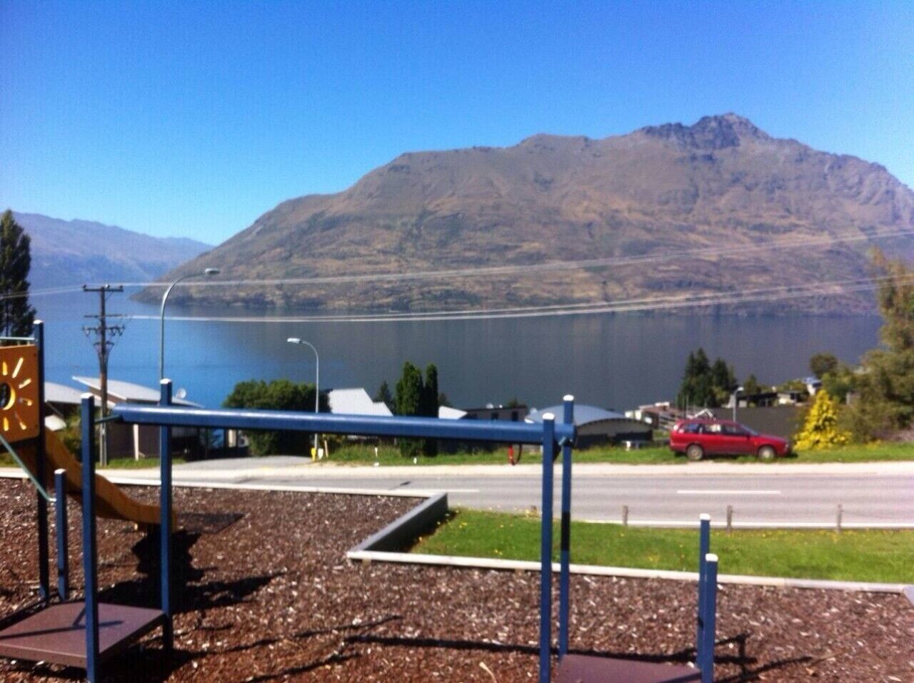 A playground with a blue swing set and a yellow climbing structure, a road with a red car, and a mountain in the background.