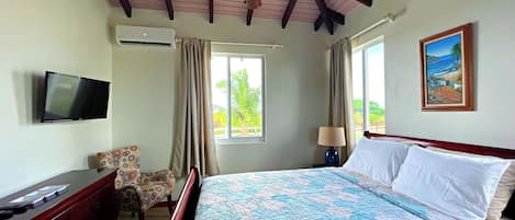 A hotel room with a bed, a TV, a ceiling fan, a window with curtains, and a painting on the wall.