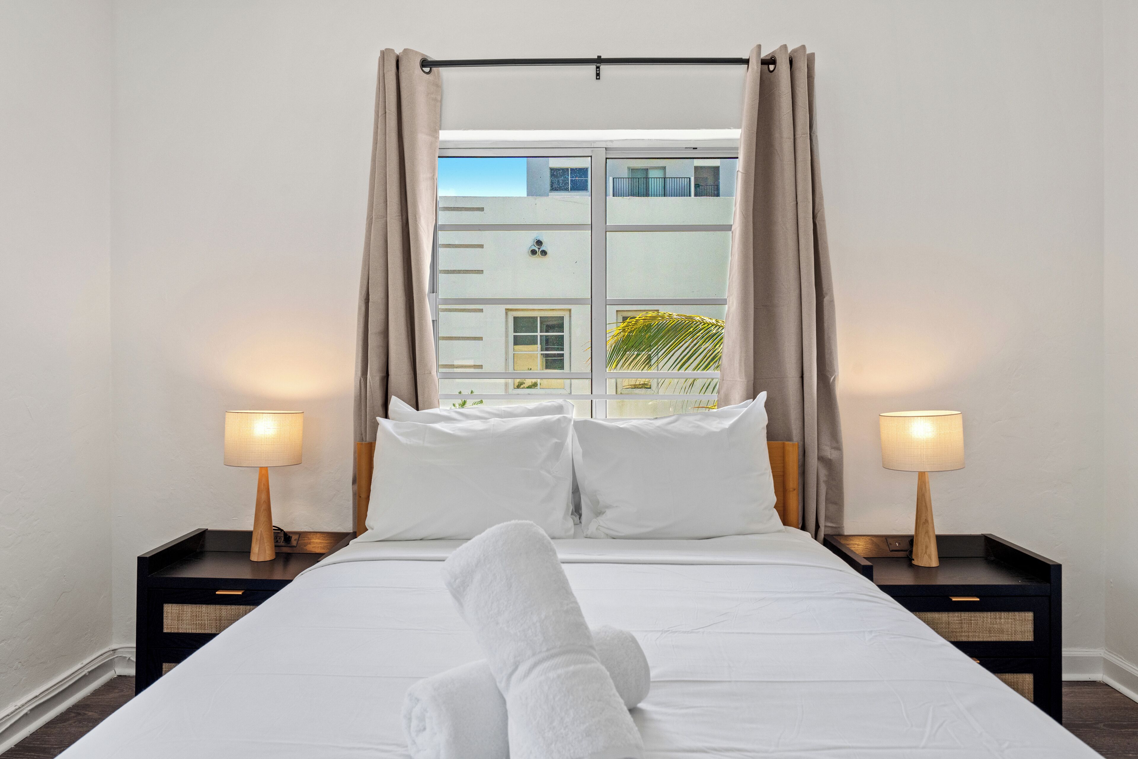 Ein Hotelzimmer mit einem Bett, zwei Nachttischen mit Lampen, einem Fenster mit Vorhängen und Blick auf Gebäude.