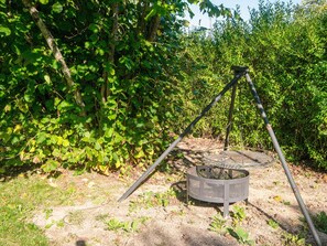 A metal grill with a circular grate, supported by a metal frame, set in a natural outdoor environment with dense green foliage in the background. - 4 Person Holiday Home in Orum Djurs (Ørum Djurs)