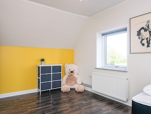 Une pièce avec un grand ours en peluche, un radiateur blanc, une fenêtre donnant sur un beau paysage et un mur jaune.