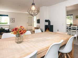 A modern dining area with a large wooden table, white chairs, a vase of flowers, a hanging light fixture, and a fireplace.