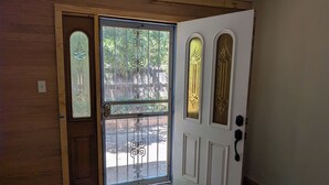Une porte en bois, dotée d’un panneau de verre et d’éléments décoratifs en métal, qui donne sur un espace extérieur avec une terrasse en bois et un paillasson jaune.
