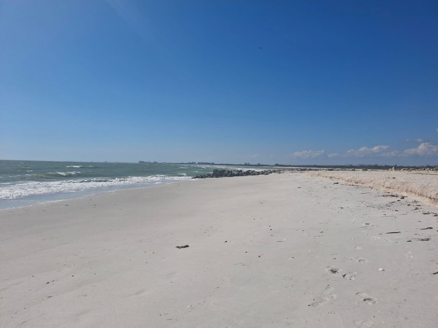 Ein weiter, sandiger Strand mit sanften Wellen, blauem Himmel und Gebäuden in der Ferne.
