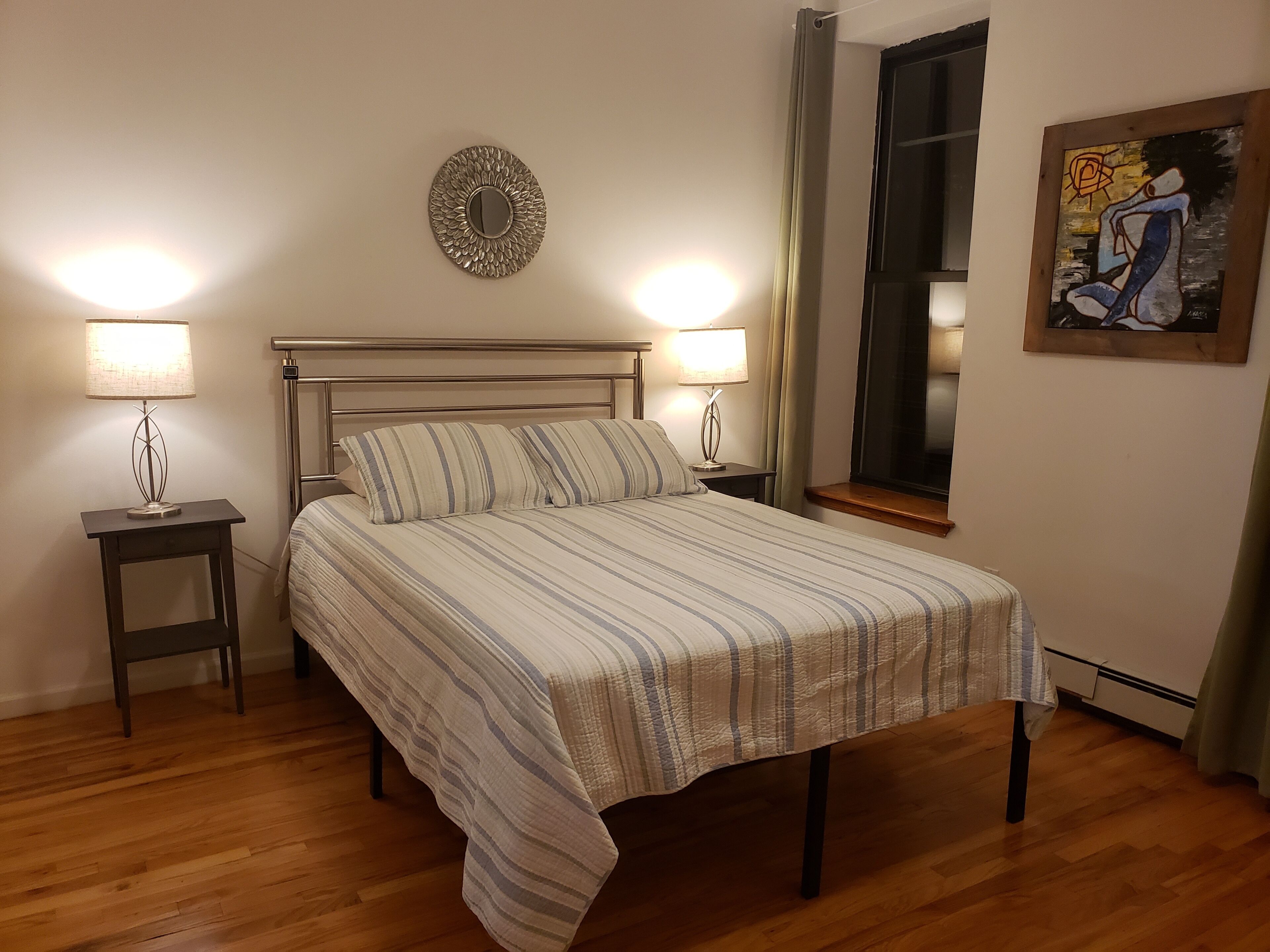 Una cama bien tendida con ropa de cama a rayas, dos mesitas de noche con lámparas, un adorno en la pared y una ventana con cortinas.