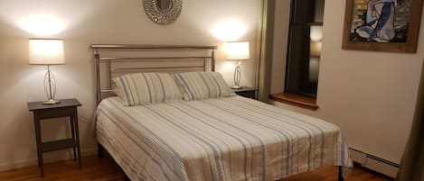 Una cama bien tendida con ropa de cama a rayas, dos mesitas de noche con lámparas, un adorno en la pared y una ventana con cortinas.
