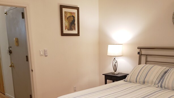 A bedroom with a bed, a nightstand, a lamp, a door, and a framed picture on the wall.